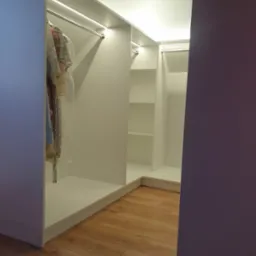 Wnętrze narożnej, białej garderoby z oświetleniem LED, widoczne ubrania na wieszakach i puste półki, parkiet na podłodze.