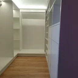 Wnętrze nowoczesnej garderoby na wymiar z białymi półkami i szafkami, oświetleniem LED oraz drewnianą podłogą, widok z perspektywy wejścia.