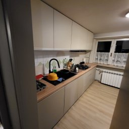 Omeblowani Mariusz Jastrzębski - Beżowa zabudowa kuchenna z drewnianym blatem, czarną armaturą i białymi kafelkami z zielonym wzorem, widok na okno z białymi firankami.