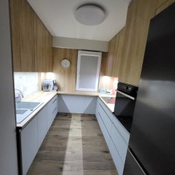 Omeblowani Mariusz Jastrzębski - Wąska kuchnia w kształcie litery U z drewnianymi szafkami górnymi i szarymi dolnymi, oświetlona lampą sufitową, z widocznym oknem i zegarem na ścianie, oraz sprzętem AGD.