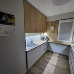 Nowoczesna kuchnia z drewnianymi szafkami górnymi i białymi dolnymi, z widocznym kalendarzem na ścianie wskazującym czerwiec 2023, zegarem ściennym i eksposem na blacie.