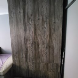 Omeblowani Mariusz Jastrzębski - Wysoka szafa z przesuwnymi drzwiami o fakturze ciemnego drewna w sypialni, widoczny fragment łóżka z fioletową pościelą