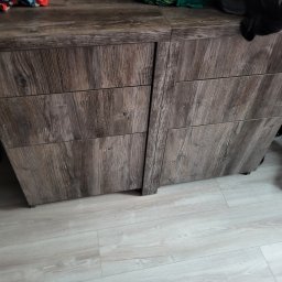 Omeblowani Mariusz Jastrzębski - Komoda z szufladami w odcieniach szarości z wyraźną strukturą drewna, widoczne niedoskonałości, ujęcie z niskiego kąta, na blacie widoczne przedmioty.