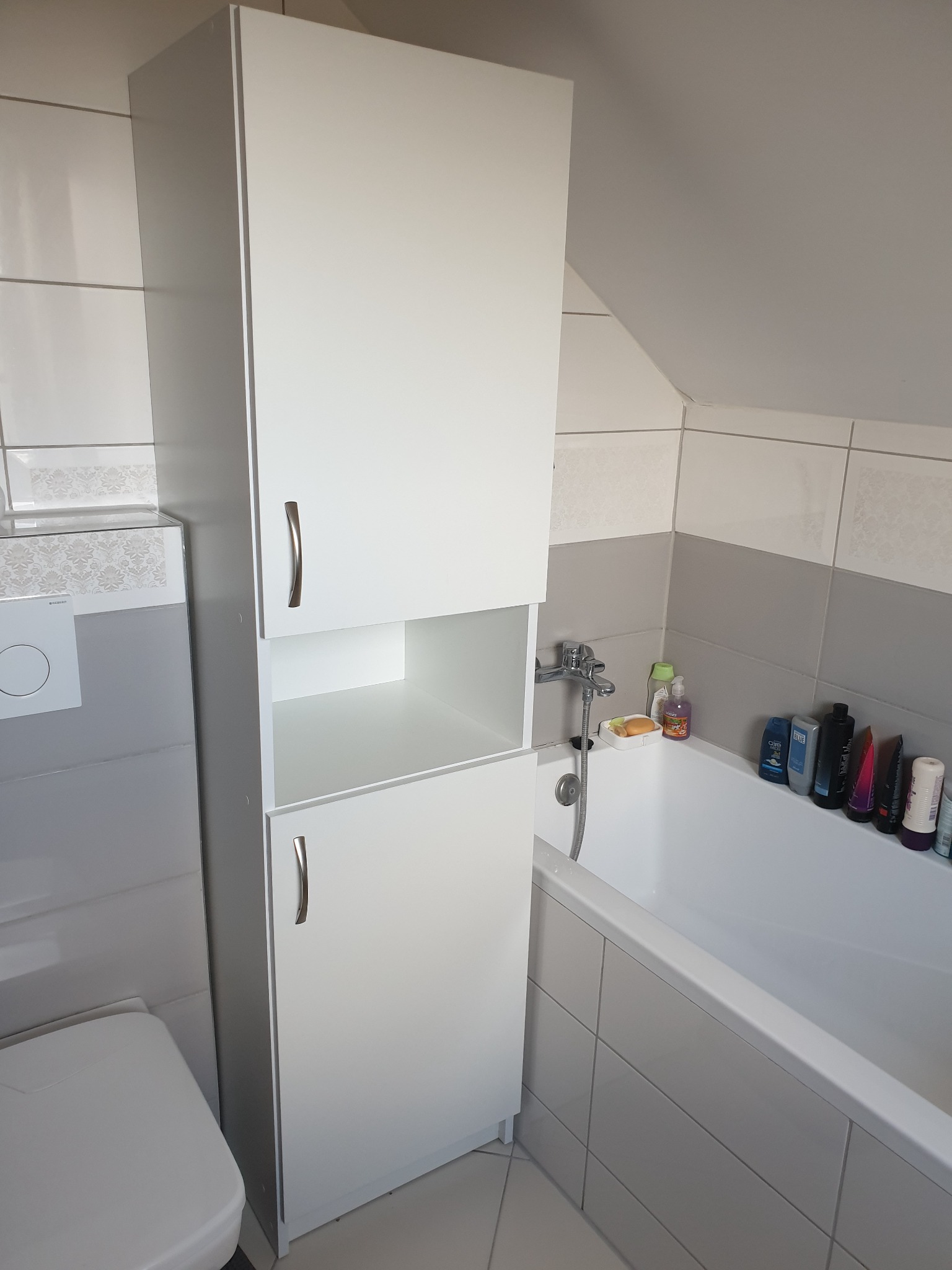 Wysoka, biała szafka łazienkowa z dwiema zamkniętymi półkami i otwartą przestrzenią pośrodku, umieszczona obok toalety i wanny w łazience z szaro-białymi płytkami.