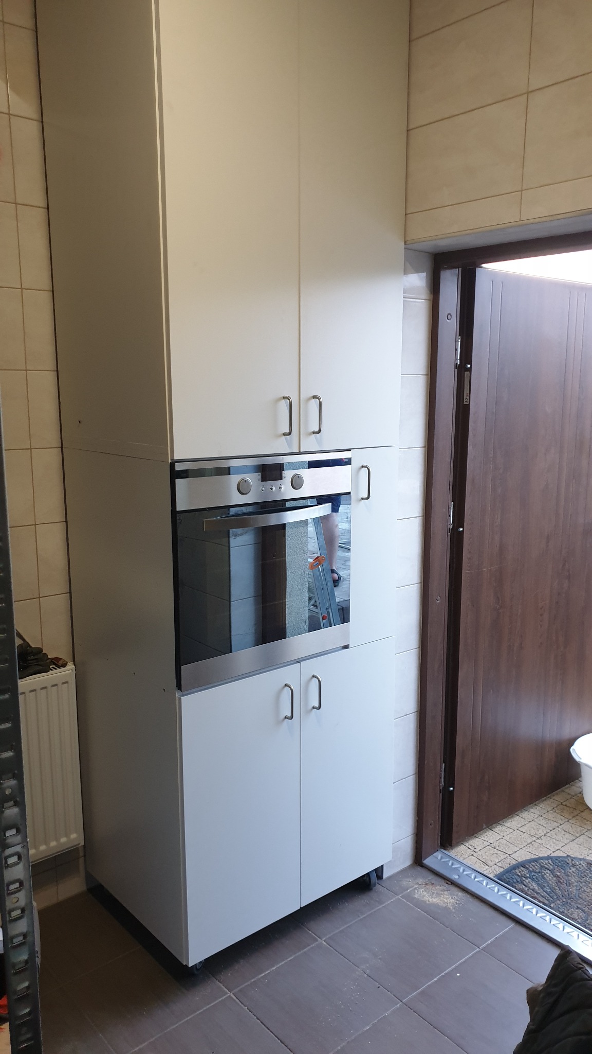 Biała zabudowa kuchenna z piekarnikiem, widoczna drabina w odbiciu szyby piekarnika, brązowe płytki podłogowe i drzwi wejściowe.