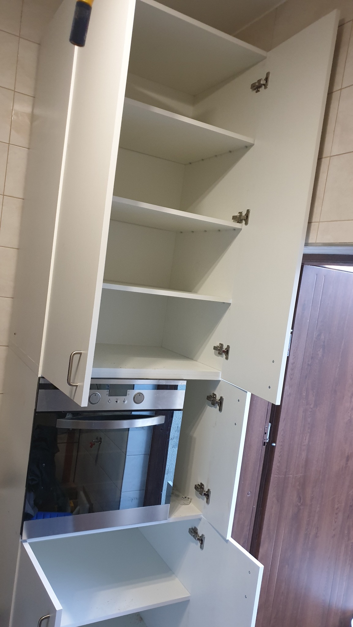 Biała szafka kuchenna z otwartymi drzwiczkami, widoczne półki, piekarnik w zabudowie, płytki na ścianie i fragment drewnianych drzwi w tle.