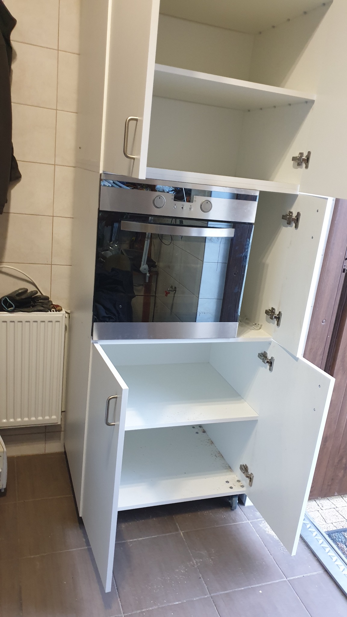 Biała zabudowa kuchenna z piekarnikiem w trakcie montażu, widoczne otwarte szafki i niedokładności montażowe, w tle grzejnik i płytki ceramiczne.