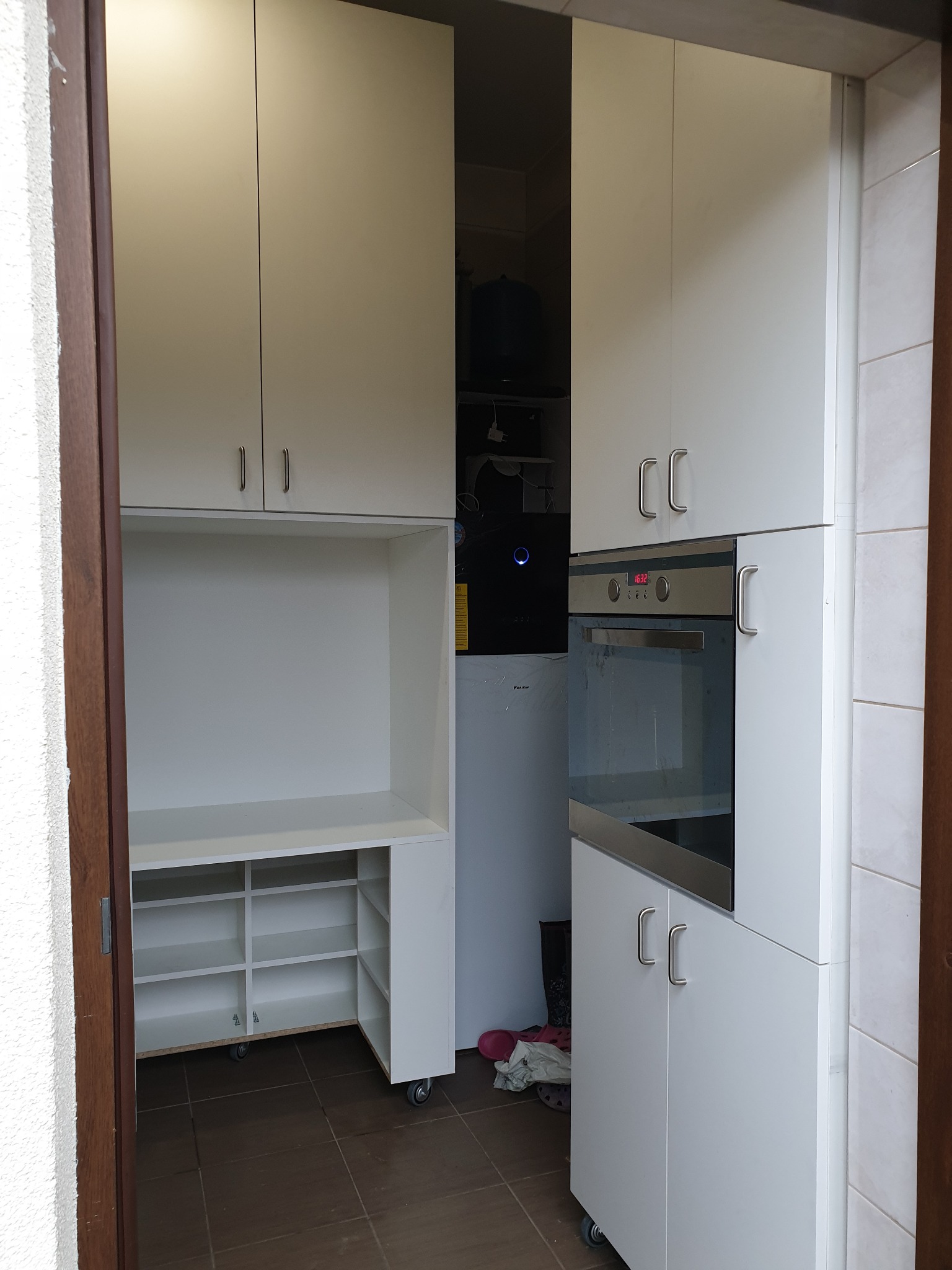 Białe szafki kuchenne z piekarnikiem i półkami na buty, widoczne przez framugę drzwi, na podłodze płytki w odcieniach brązu.