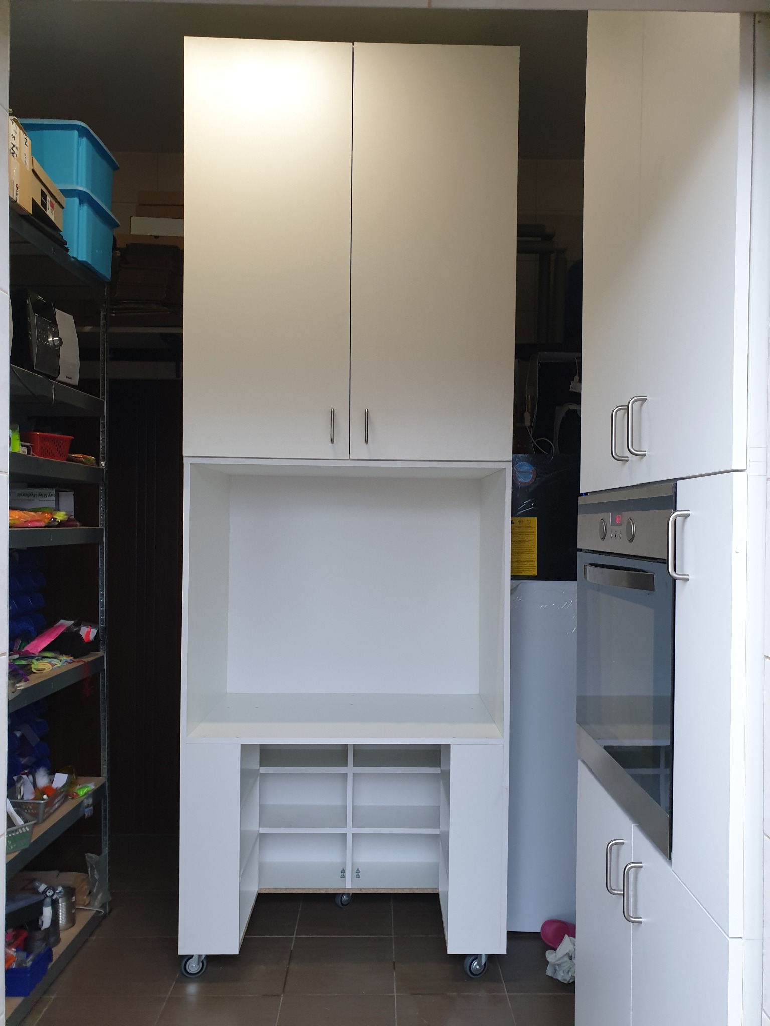 Biała zabudowa kuchenna z szafkami górnymi i otwartą przestrzenią roboczą na kółkach, widoczna na tle półek z różnymi przedmiotami, obok piekarnika w zabudowie.
