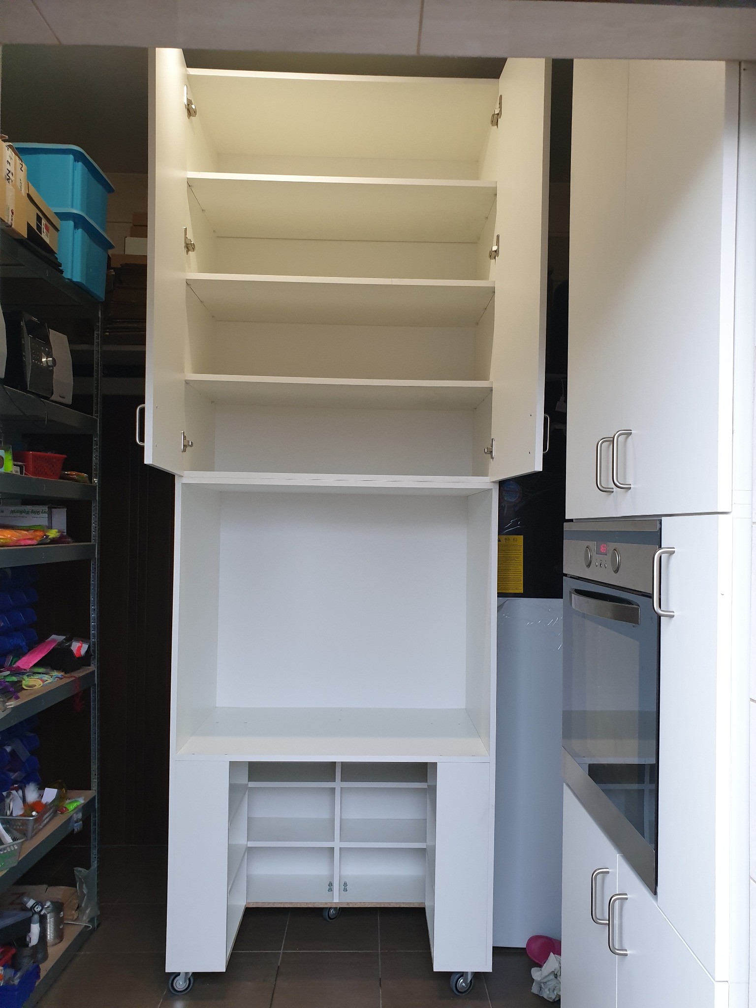 Biała, modułowa szafka z otwartymi górnymi drzwiczkami, widoczne półki wewnątrz. Dolna część szafki na kółkach, z otwartymi przegródkami. Obok zabudowa kuchenna z piekarnikiem.