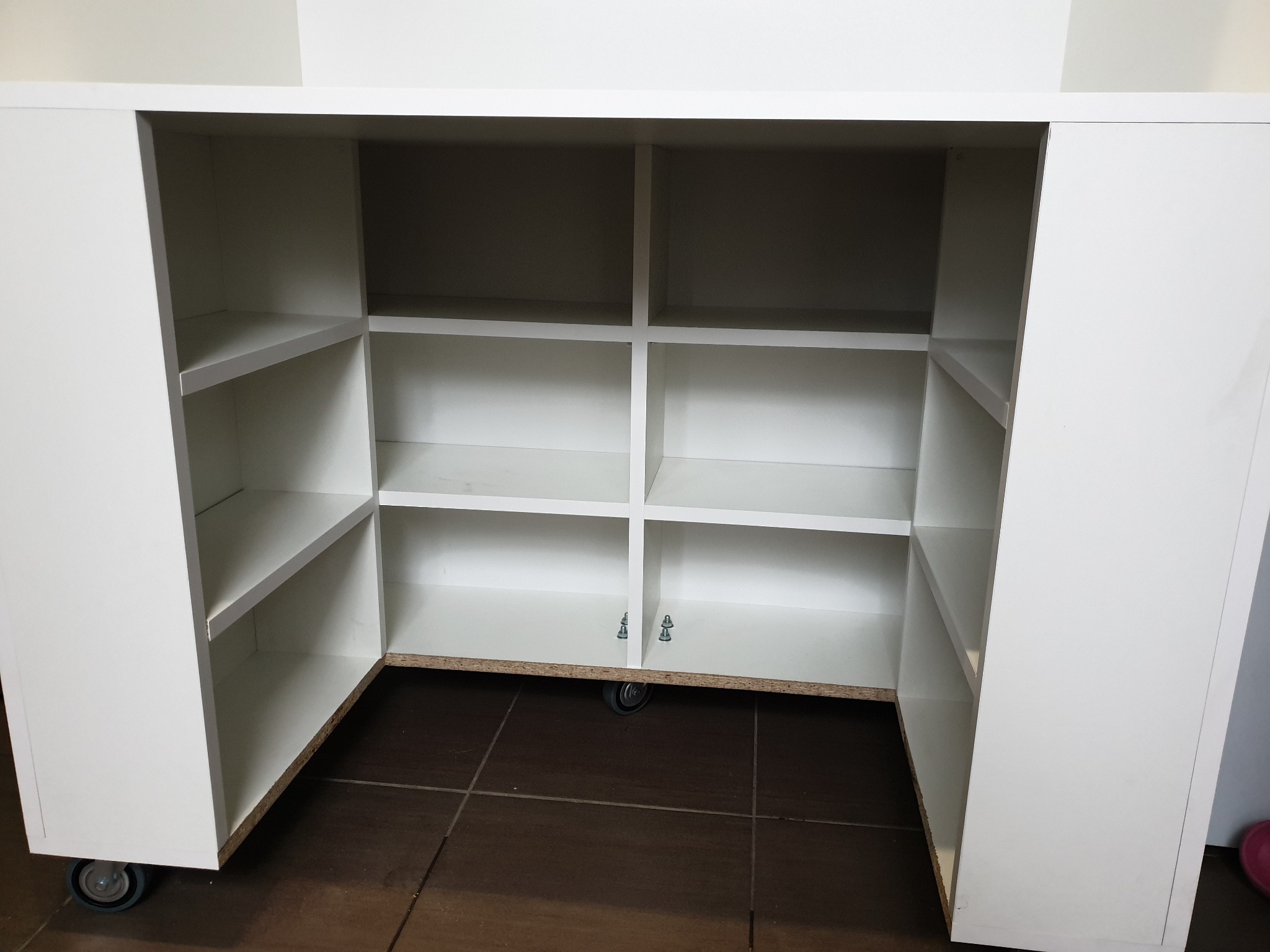 Biała, modułowa szafka z półkami i kółkami, widoczne mocowania półek, na brązowej podłodze z płytek.