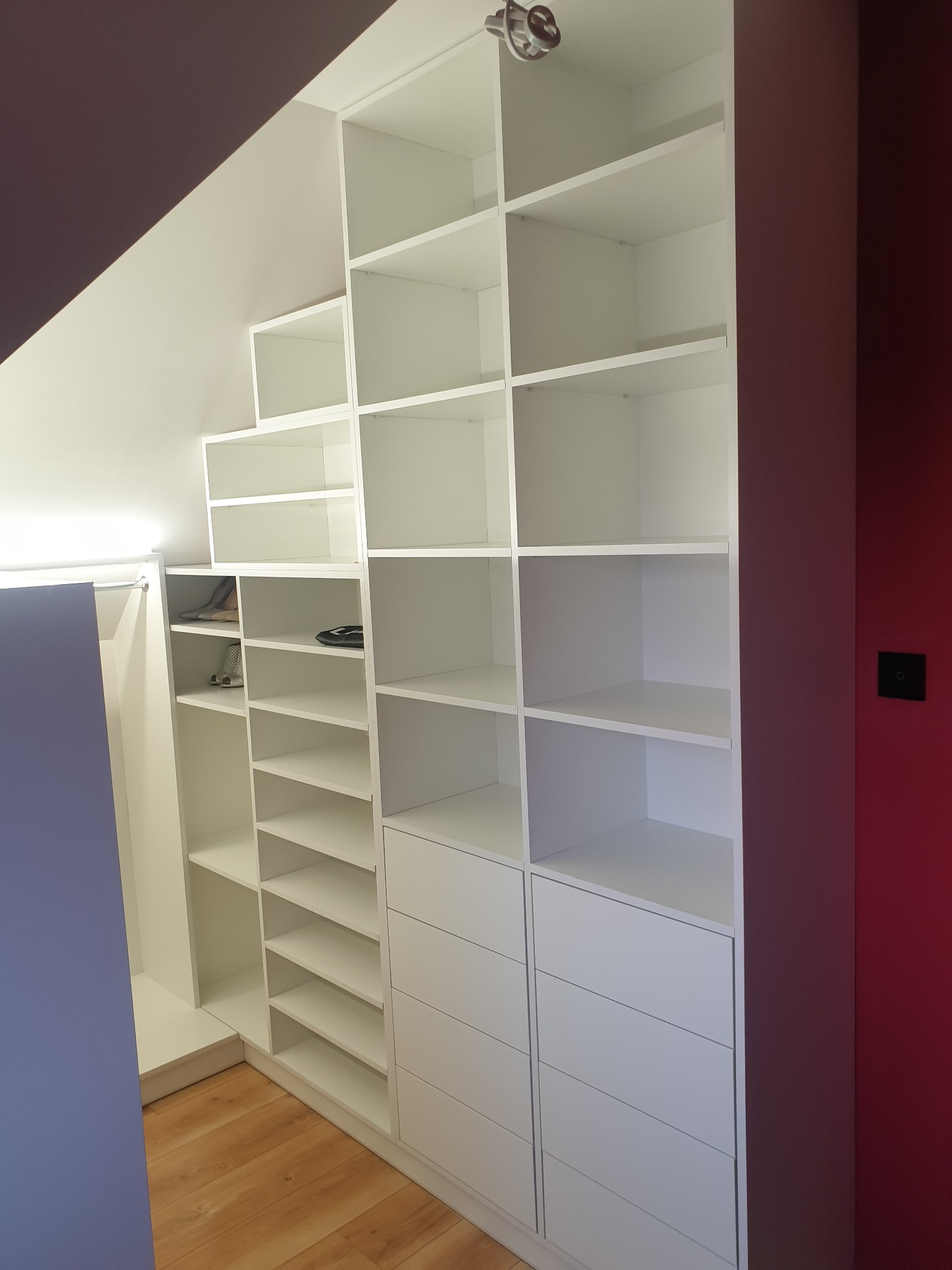 Biała zabudowa szafy wnękowej na poddaszu ze skosami, z półkami i szufladami, oświetlenie punktowe na suficie.