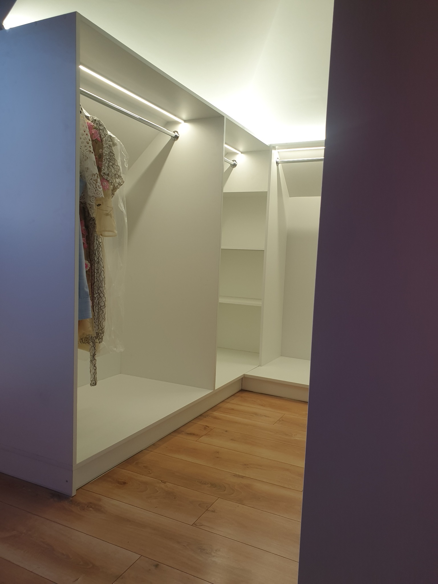 Wnętrze narożnej, białej garderoby z oświetleniem LED, widoczne ubrania na wieszakach i puste półki, parkiet na podłodze.