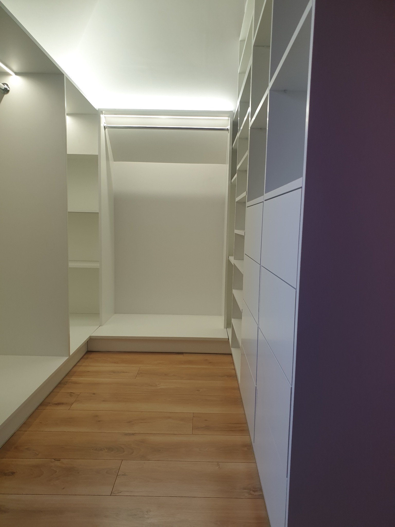 Wnętrze nowoczesnej garderoby na wymiar z białymi półkami i szafkami, oświetleniem LED oraz drewnianą podłogą, widok z perspektywy wejścia.