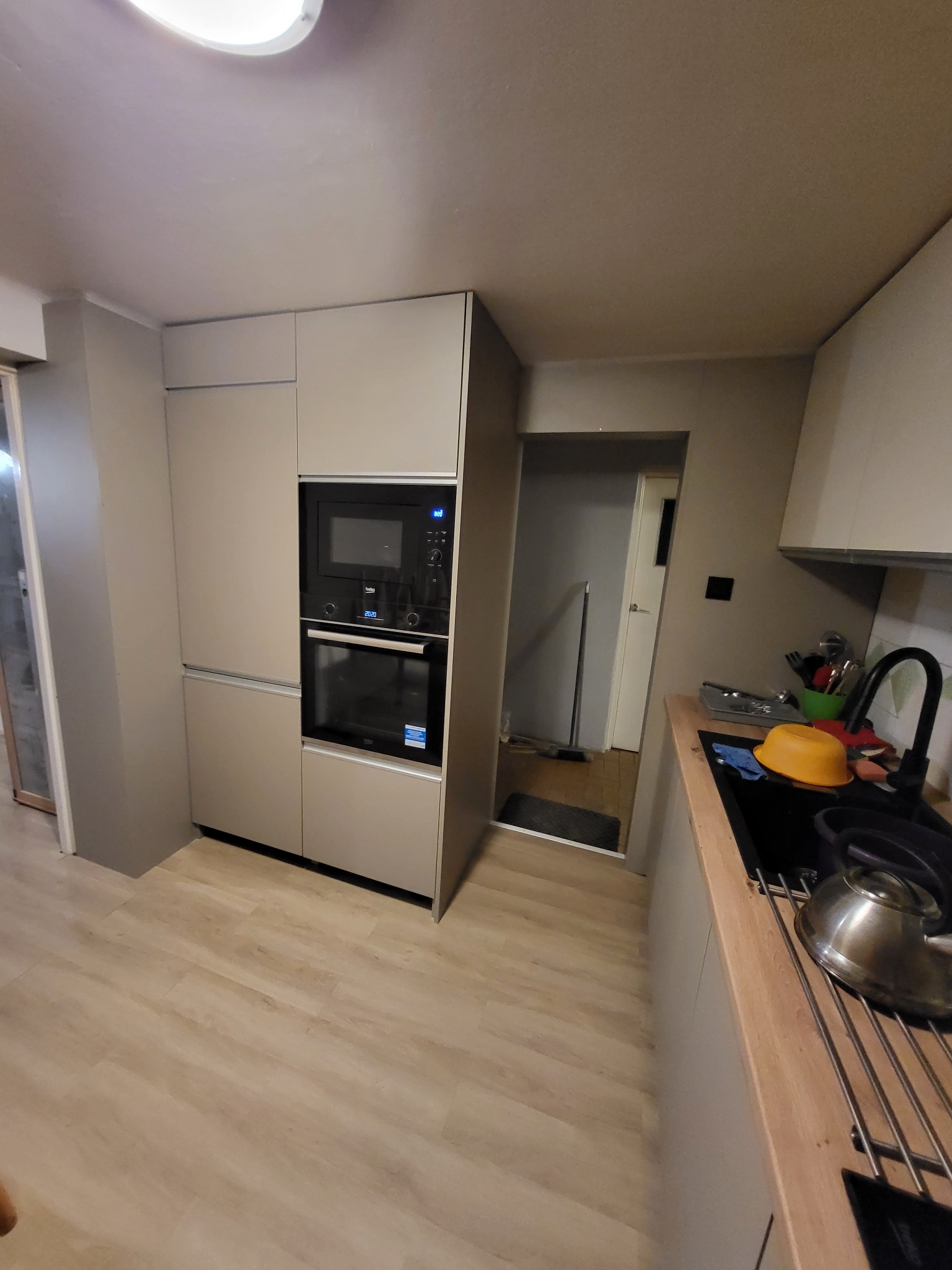 Szare, minimalistyczne meble kuchenne z wbudowanym piekarnikiem i mikrofalówką; widoczny fragment blatu z czarnym zlewem i czajnikiem.