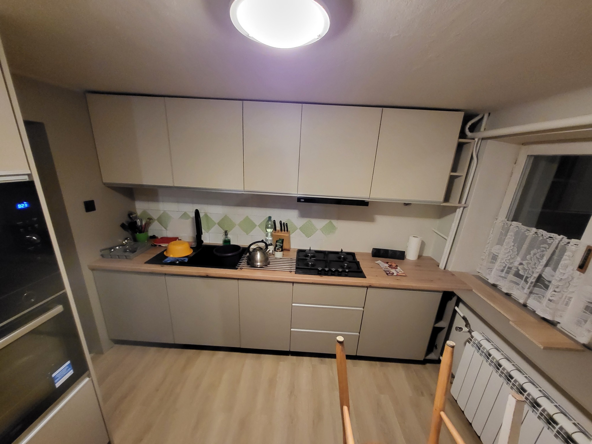 Jasna kuchnia z beżowymi frontami szafek, czarnym blatem i płytą grzewczą, widok na okno z białymi firankami i grzejnik.