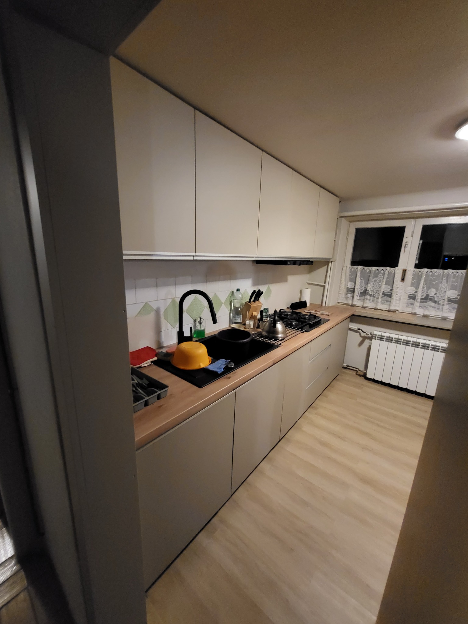 Beżowa zabudowa kuchenna z drewnianym blatem, czarną armaturą i białymi kafelkami z zielonym wzorem, widok na okno z białymi firankami.