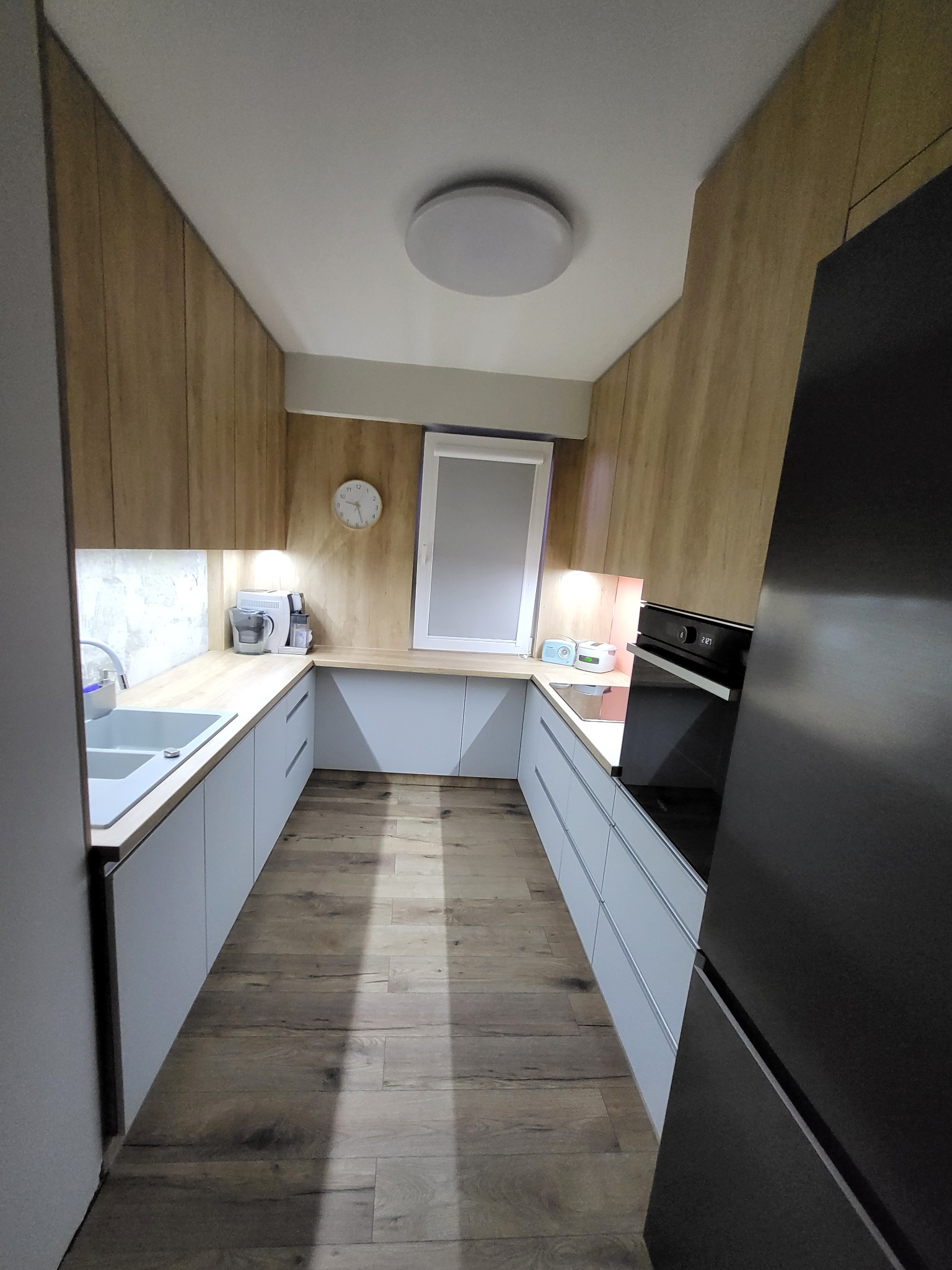 Wąska kuchnia w kształcie litery U z drewnianymi szafkami górnymi i szarymi dolnymi, oświetlona lampą sufitową, z widocznym oknem i zegarem na ścianie, oraz sprzętem AGD.