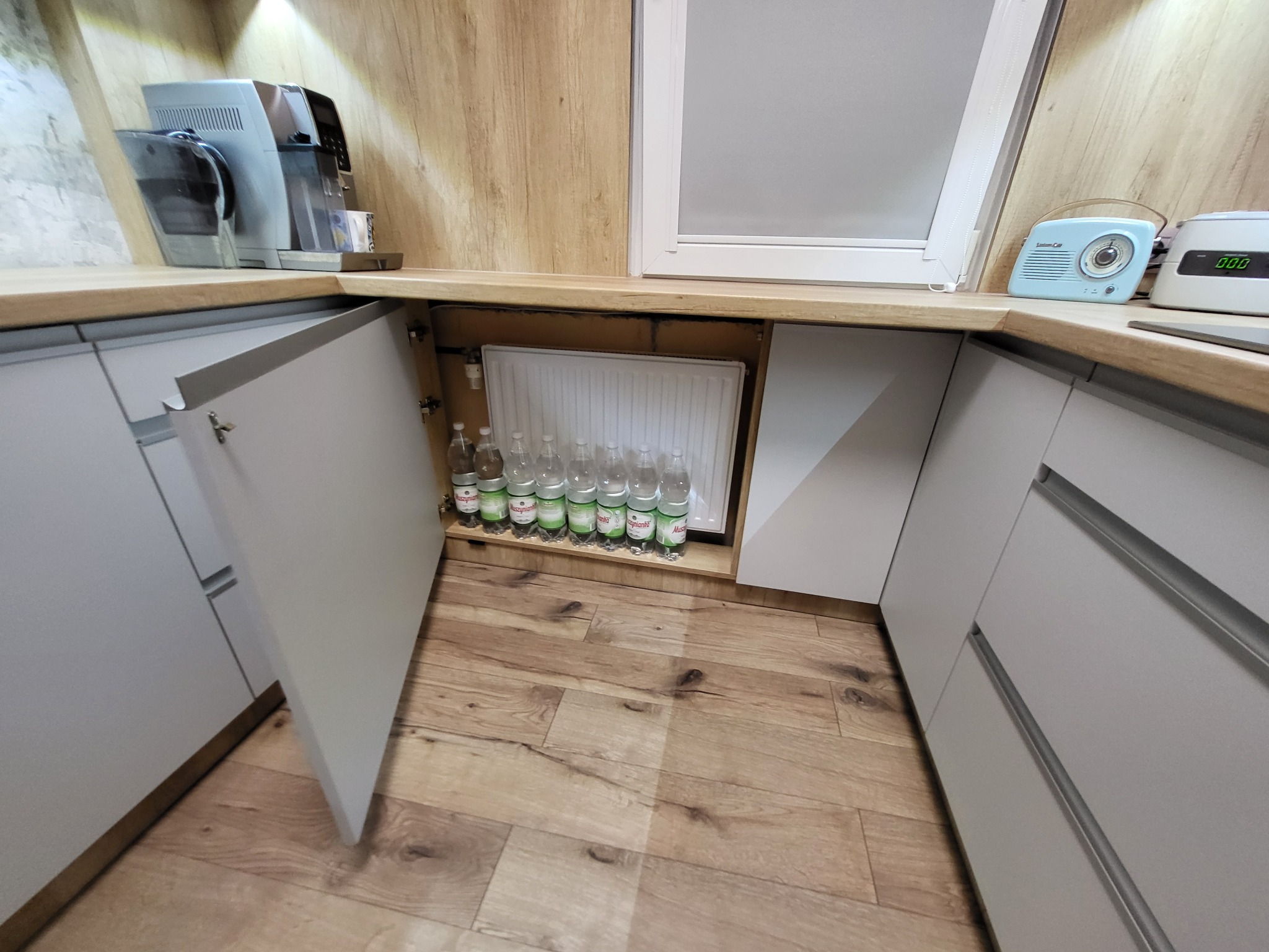 Szara zabudowa kuchenna z otwartą szafką pod blatem, w której widać grzejnik i zapas butelek wody mineralnej. Na blacie ekspres do kawy i radio w stylu retro.