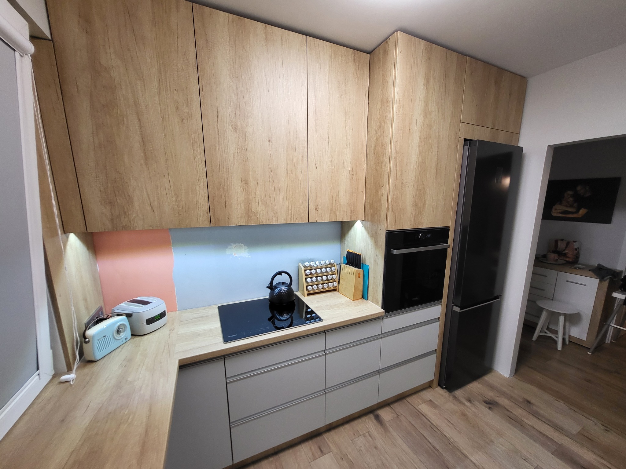 Nowoczesna zabudowa kuchenna z drewnianymi frontami szafek górnych i szarymi dolnymi w Sieradzu, z czarnym piekarnikiem i lodówką, uzupełniona o czajnik na płycie indukcyjnej i akcesoria kuchenne.