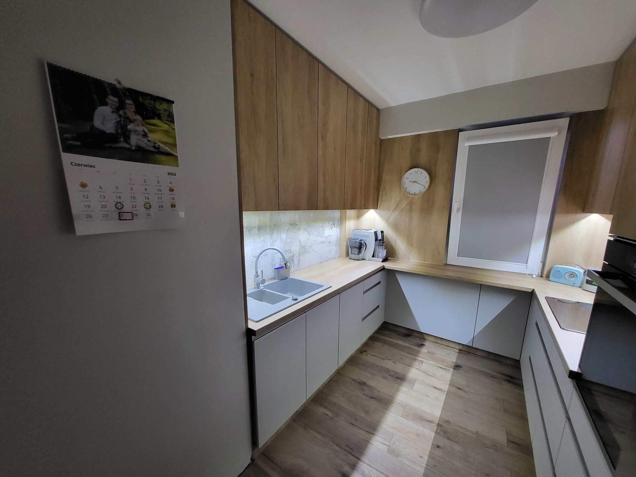 Nowoczesna kuchnia z drewnianymi szafkami górnymi i białymi dolnymi, z widocznym kalendarzem na ścianie wskazującym czerwiec 2023, zegarem ściennym i eksposem na blacie.