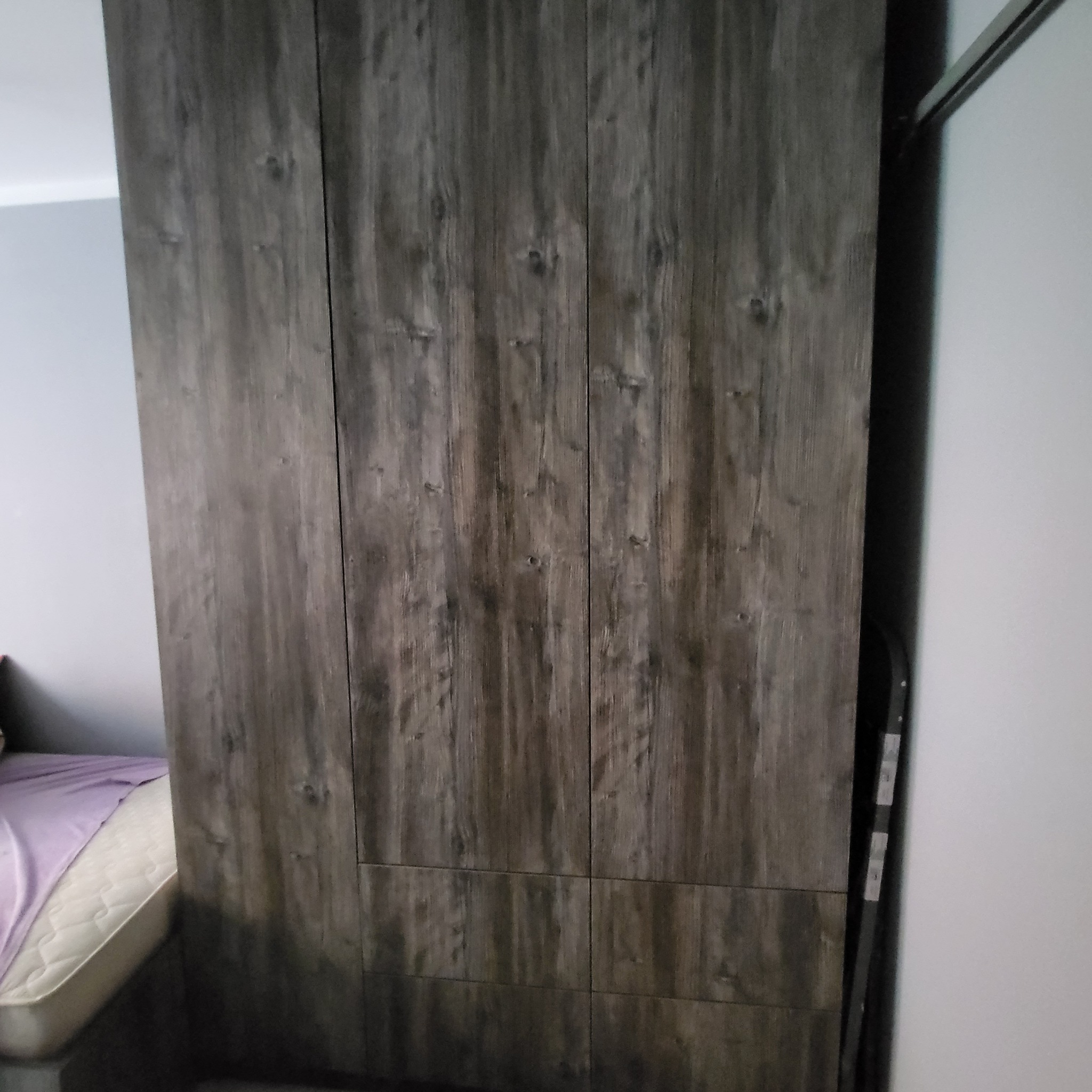 Wysoka szafa z przesuwnymi drzwiami o fakturze ciemnego drewna w sypialni, widoczny fragment łóżka z fioletową pościelą