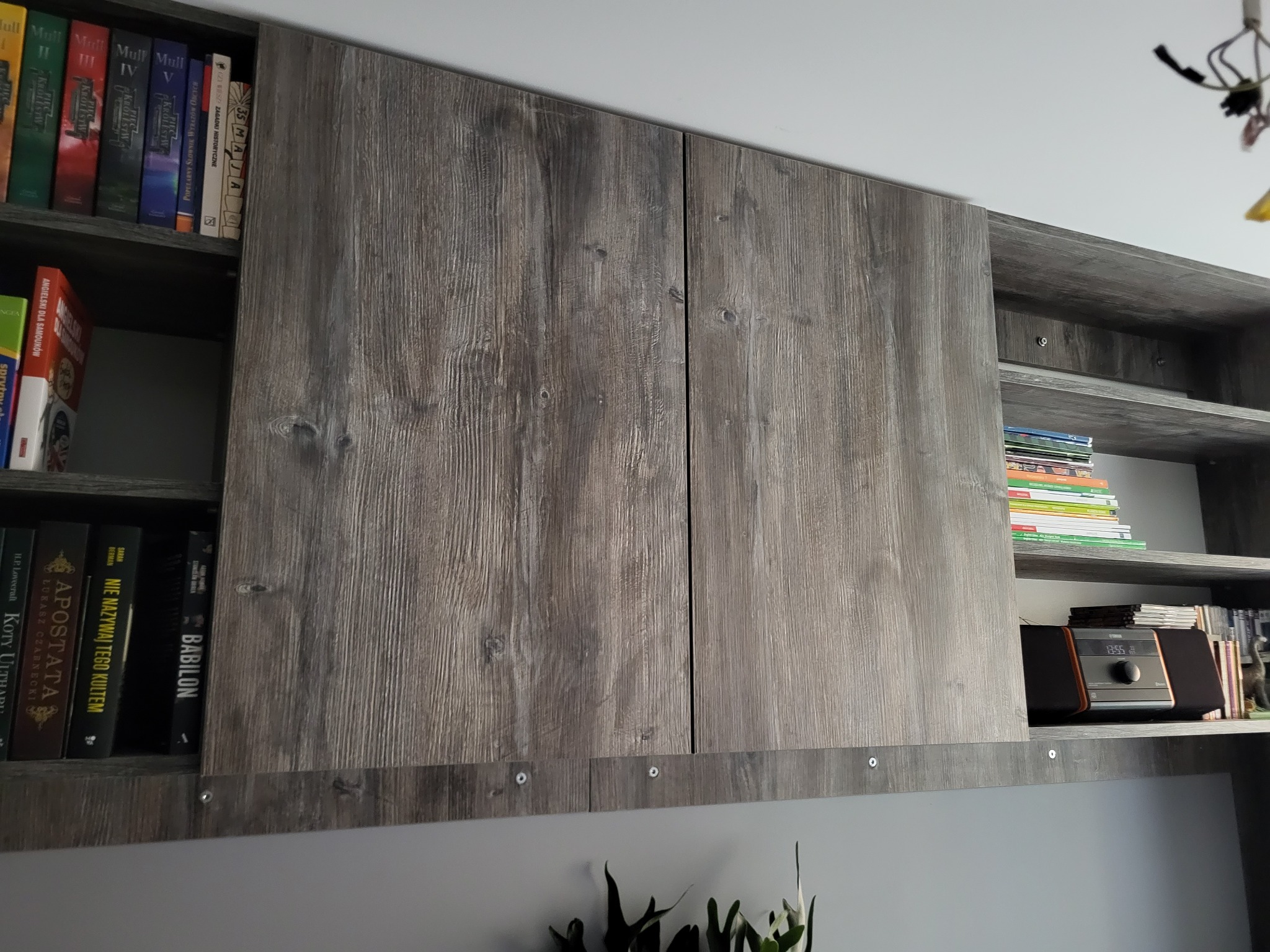 Szare drewniane szafki wiszące z książkami i radiem na półkach, widoczne okucia mocujące.