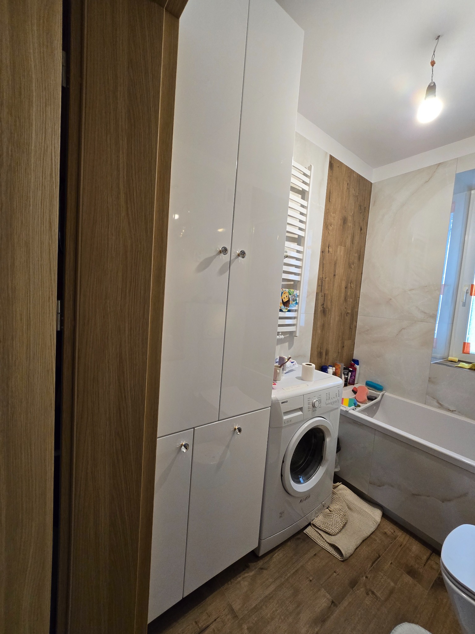 Wysoka, biała szafa łazienkowa z połyskiem, z kryształowymi gałkami, stojąca obok pralki w nowoczesnej łazience z drewnianymi i marmurowymi akcentami.