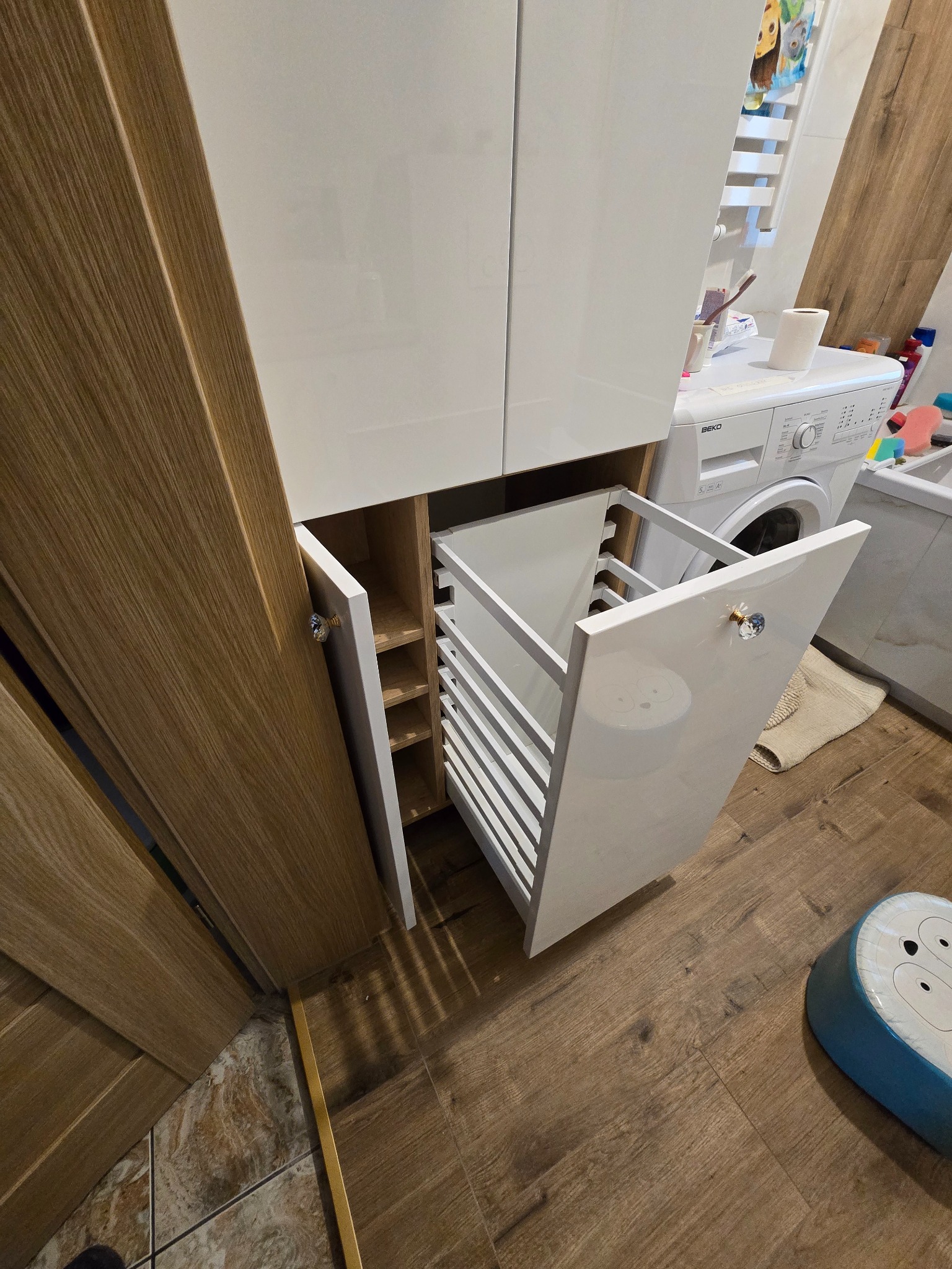 Biała szafa łazienkowa na wymiar z wysuwanym koszem na pranie, kryształowe gałki, drewnopodobna okleina frontów, pralka w tle.