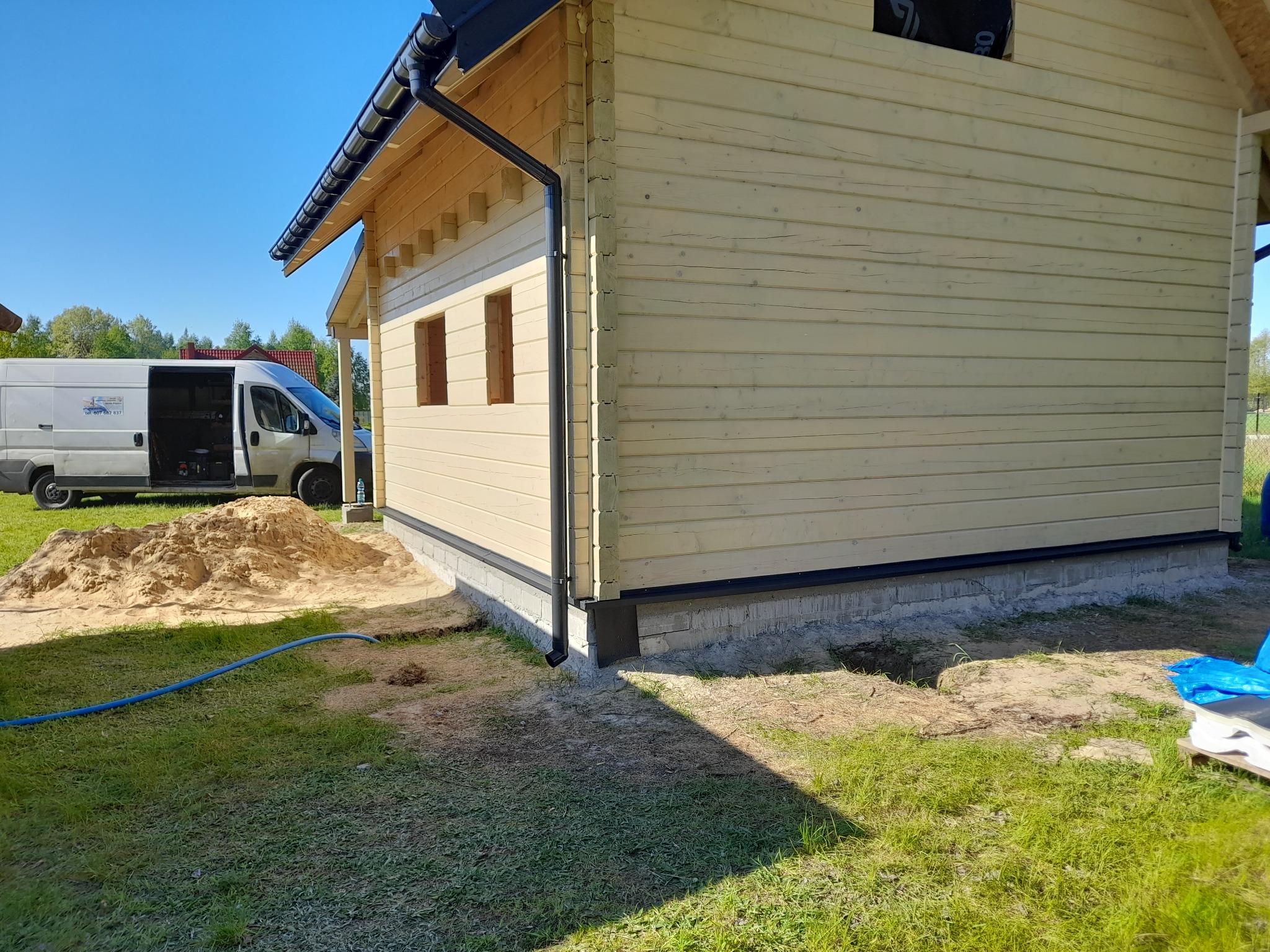 Surowy stan budowy domu z drewnianych bali, widoczne elementy konstrukcyjne, orynnowanie, fundament, hałda piasku i samochód dostawczy w tle.