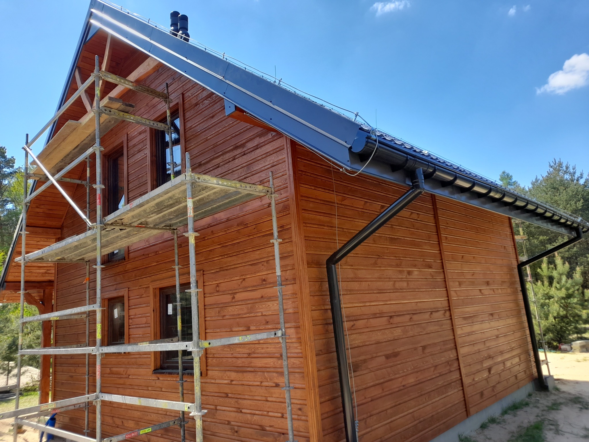 Wznoszenie domu o drewnianej elewacji z widocznymi rusztowaniami i czarnymi rynnami na tle błękitnego nieba.
