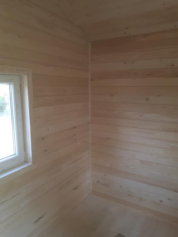 Wnętrze surowego drewnianego domku szkieletowego z widocznym oknem, ukazujące konstrukcję ścian z desek boazeryjnych przed wykończeniem.