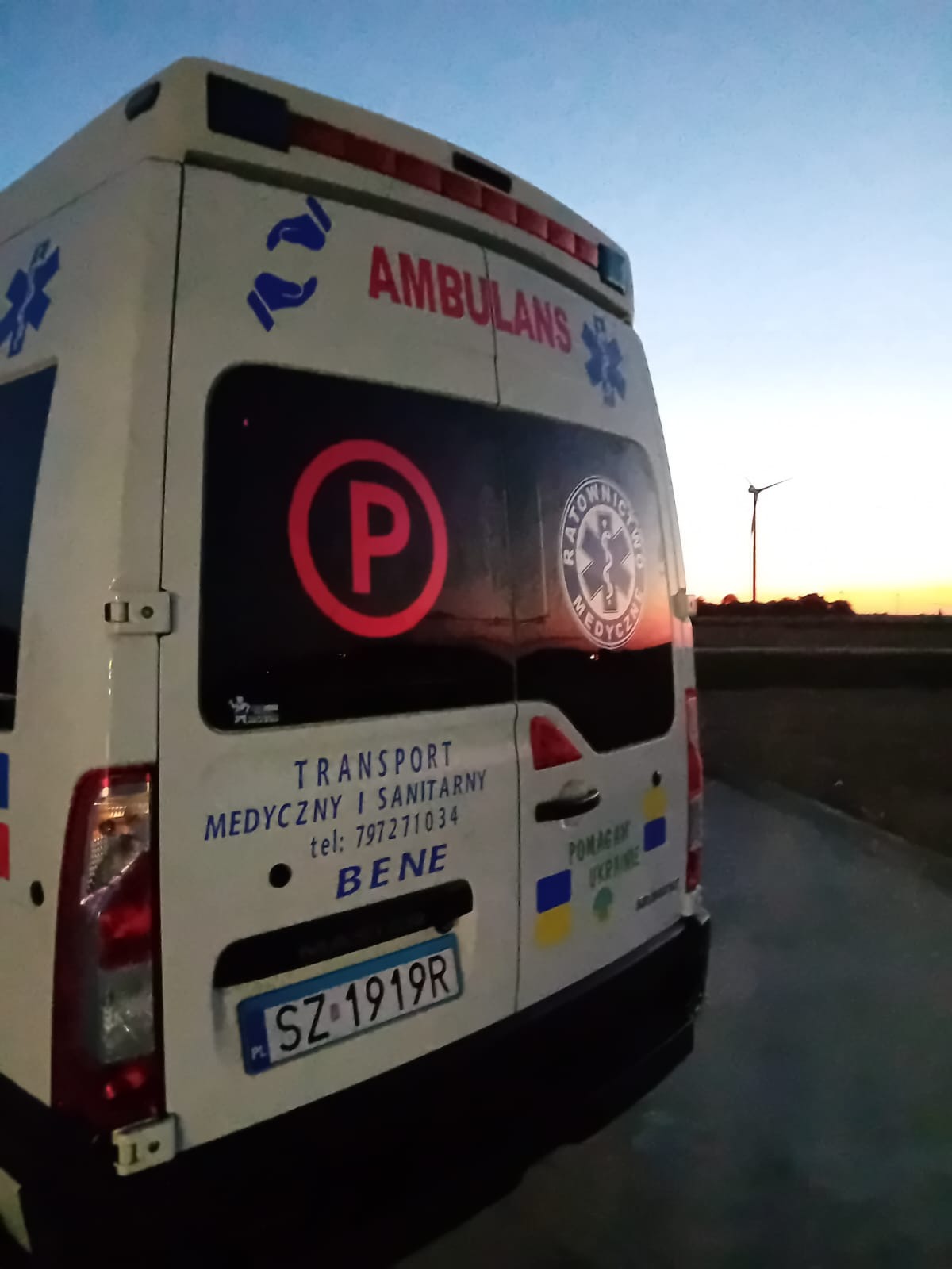 Tył ambulansu medycznego z widocznym znakiem 'P' na szybie, logo firmy 'Ratownictwo Medyczne' oraz napisem 'Transport Medyczny i Sanitarny' z numerem telefonu, flaga Ukrainy i w tle zachodzące...