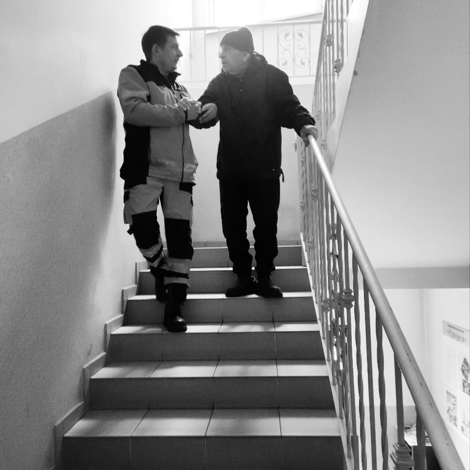 Opiekun w kombinezonie pomaga starszemu mężczyźnie w czapce zejść po schodach, wspierając go za rękę przy balustradzie.