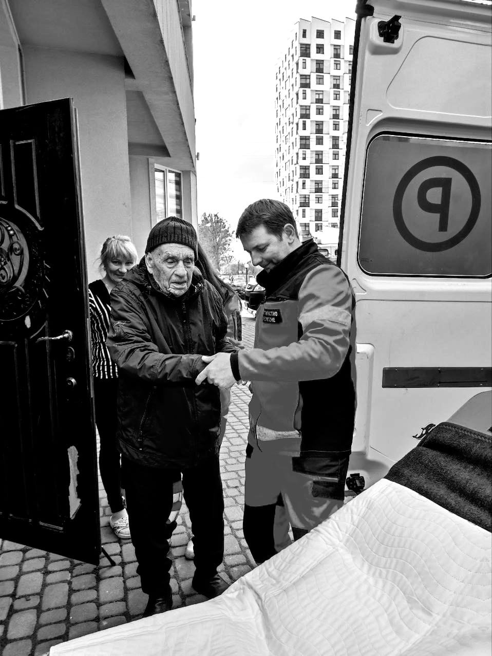 Ratownik medyczny pomaga starszemu mężczyźnie wsiąść do ambulansu na tle bloku mieszkalnego, kobieta obserwuje zza drzwi budynku.