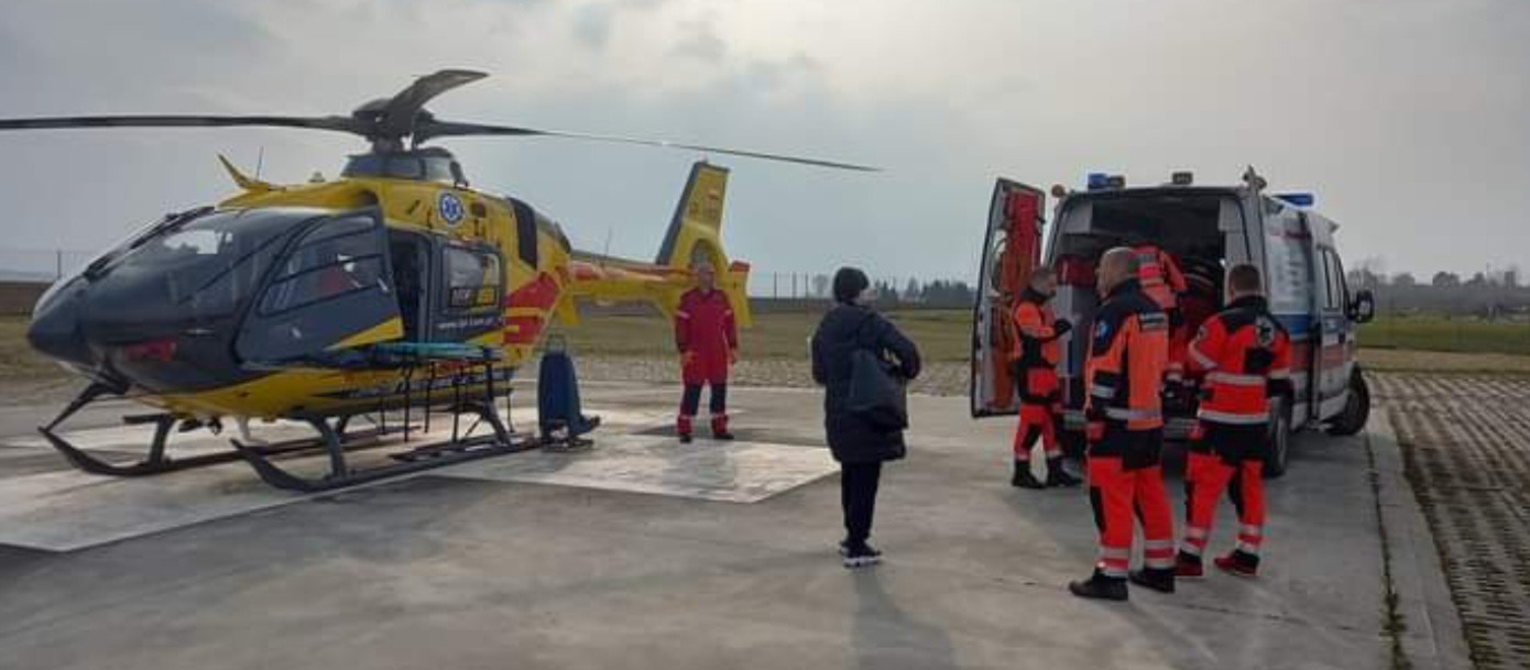 Żółto-czerwony helikopter Lotniczego Pogotowia Ratunkowego na płycie lotniska, obok ambulansu i zespołu ratowników medycznych w pomarańczowych kombinezonach, w tle krajobraz polny pod pochmurnym...