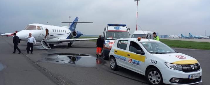 Obsługa pasażerów na lotnisku: biały prywatny odrzutowiec z otwartymi drzwiami i wysuniętymi schodami, obok ambulans i samochód lotniskowy z logo LS Airport Services na płycie lotniska.
