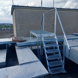 F.H.U. Tomspark - Metalowa konstrukcja schodów z platformą na dachu budynku, widok na miasto w tle.