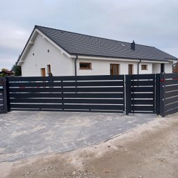 F.H.U. Tomspark - Nowoczesna, przesuwna brama wjazdowa z poziomych, grafitowych profili, na tle białego domu z ciemną dachówką i brukowanym podjazdem.