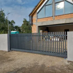 Nowoczesna, przesuwna, metalowa brama w kolorze grafitowym, z pionowymi szczeblami, zamontowana przed surowym budynkiem w trakcie budowy, z widocznymi pustakami i oknami.