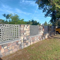 F.H.U. Tomspark - Kamienno-metalowe ogrodzenie z ażurowym wzorem, widziane z perspektywy trawnika w słoneczny dzień. Nietypowy design paneli metalowych.