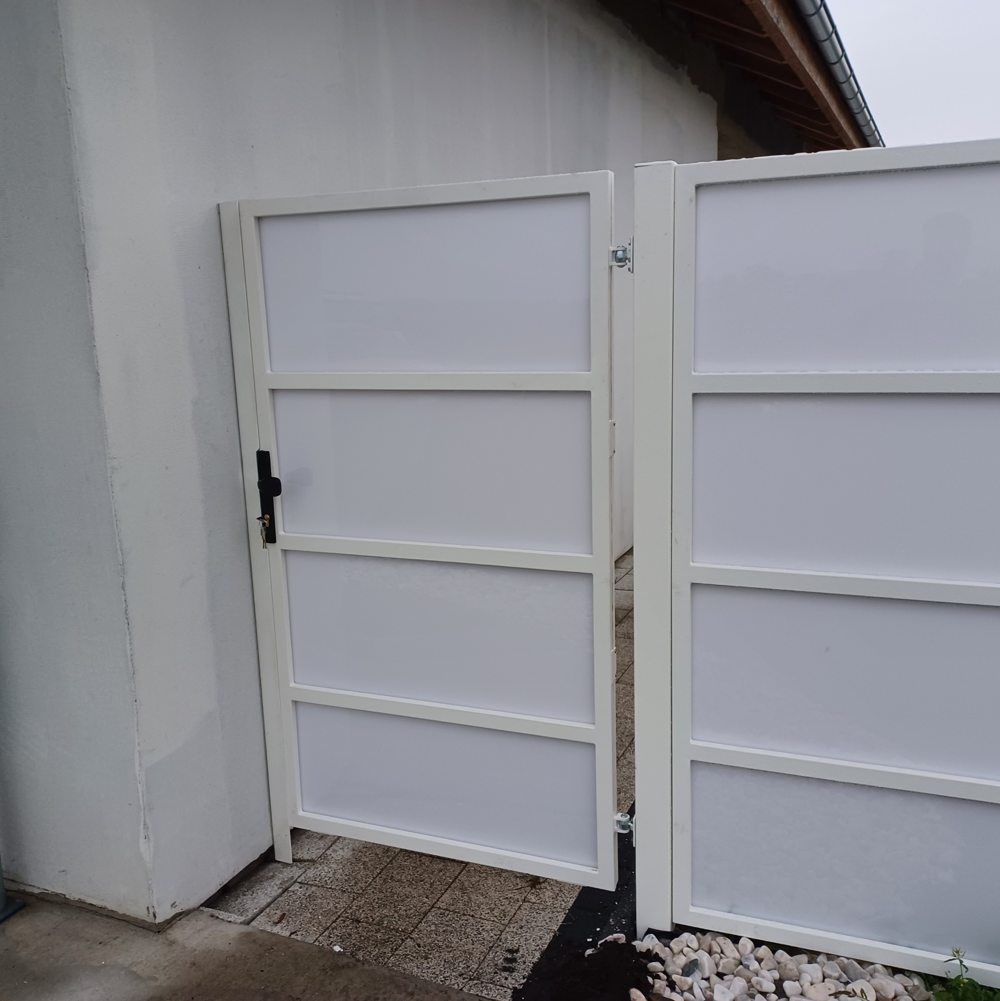 Otwarta, biała brama wjazdowa z wypełnieniem, widoczny zamek i klucze, zamontowana przy szarym, otynkowanym budynku.