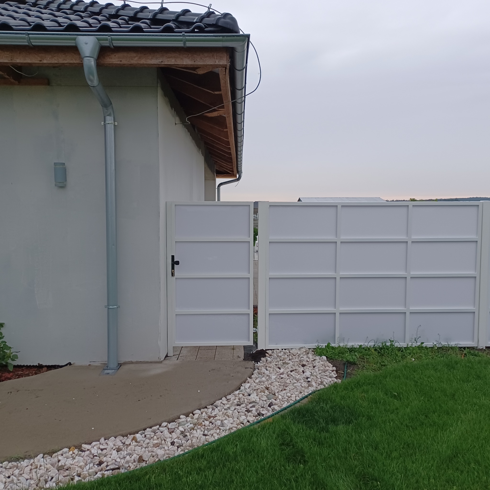 Nowoczesna, biała brama wjazdowa z metalową konstrukcją, częściowo otwarta, z widocznym fragmentem domu i trawnikiem w tle.