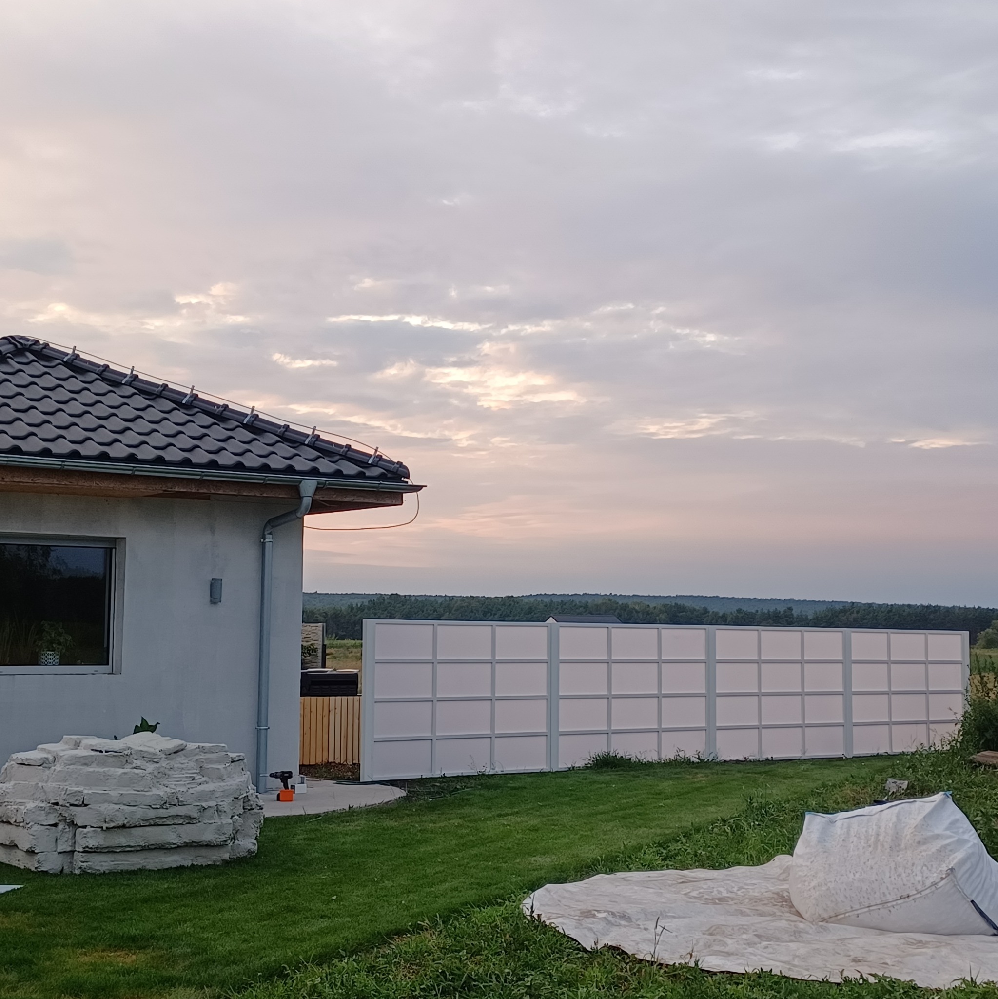 Nowoczesne ogrodzenie panelowe w kolorze białym, widoczne zza rogu domu z ciemną dachówką, na tle zachodzącego słońca i zielonego trawnika z rozsypanymi materiałami budowlanymi.