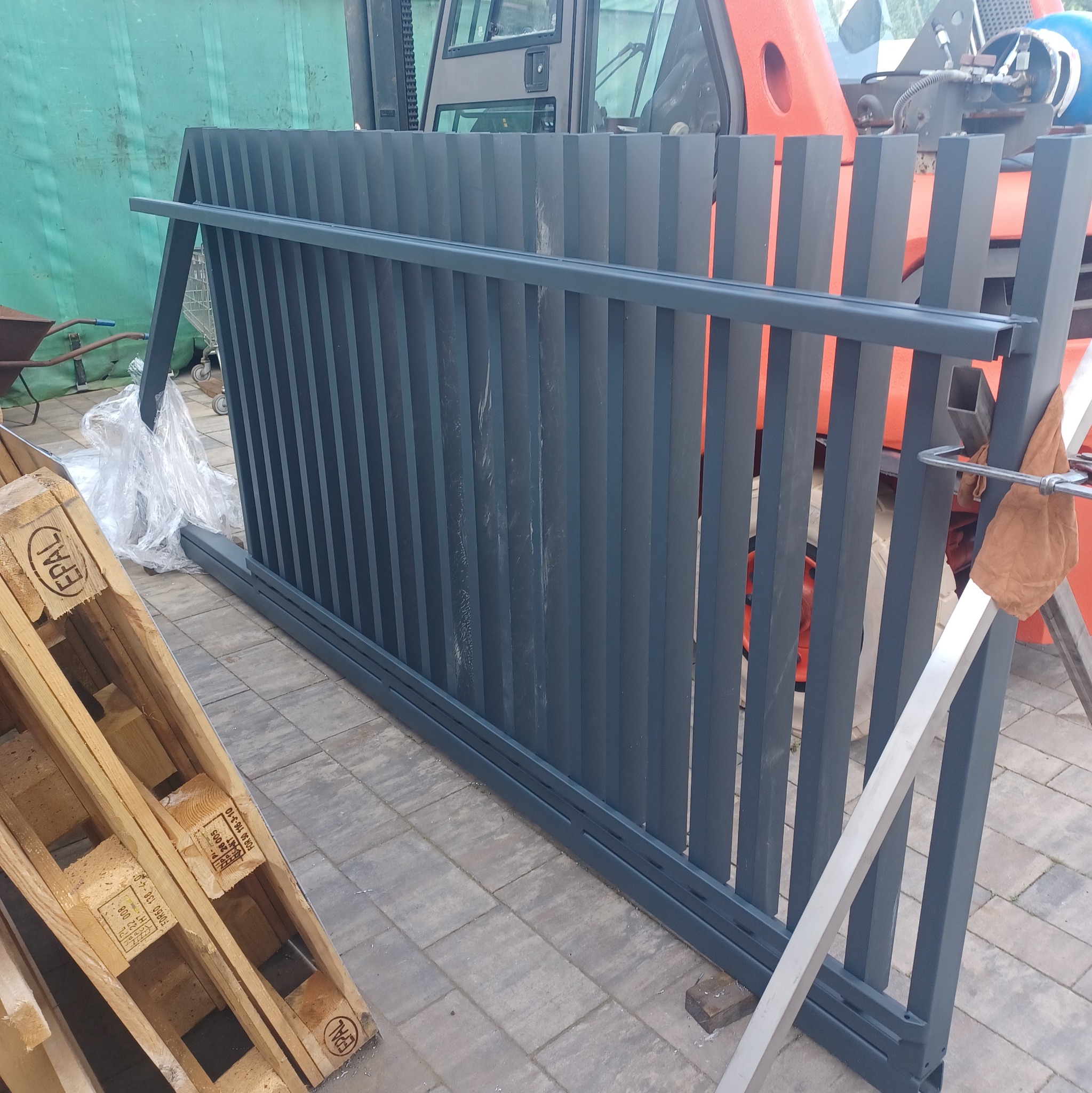 Fragment metalowego ogrodzenia w kolorze grafitowym, o pionowych szczeblach, oparty o paletę i podparty metalowym profilem, na tle wózka widłowego i zielonej plandeki.