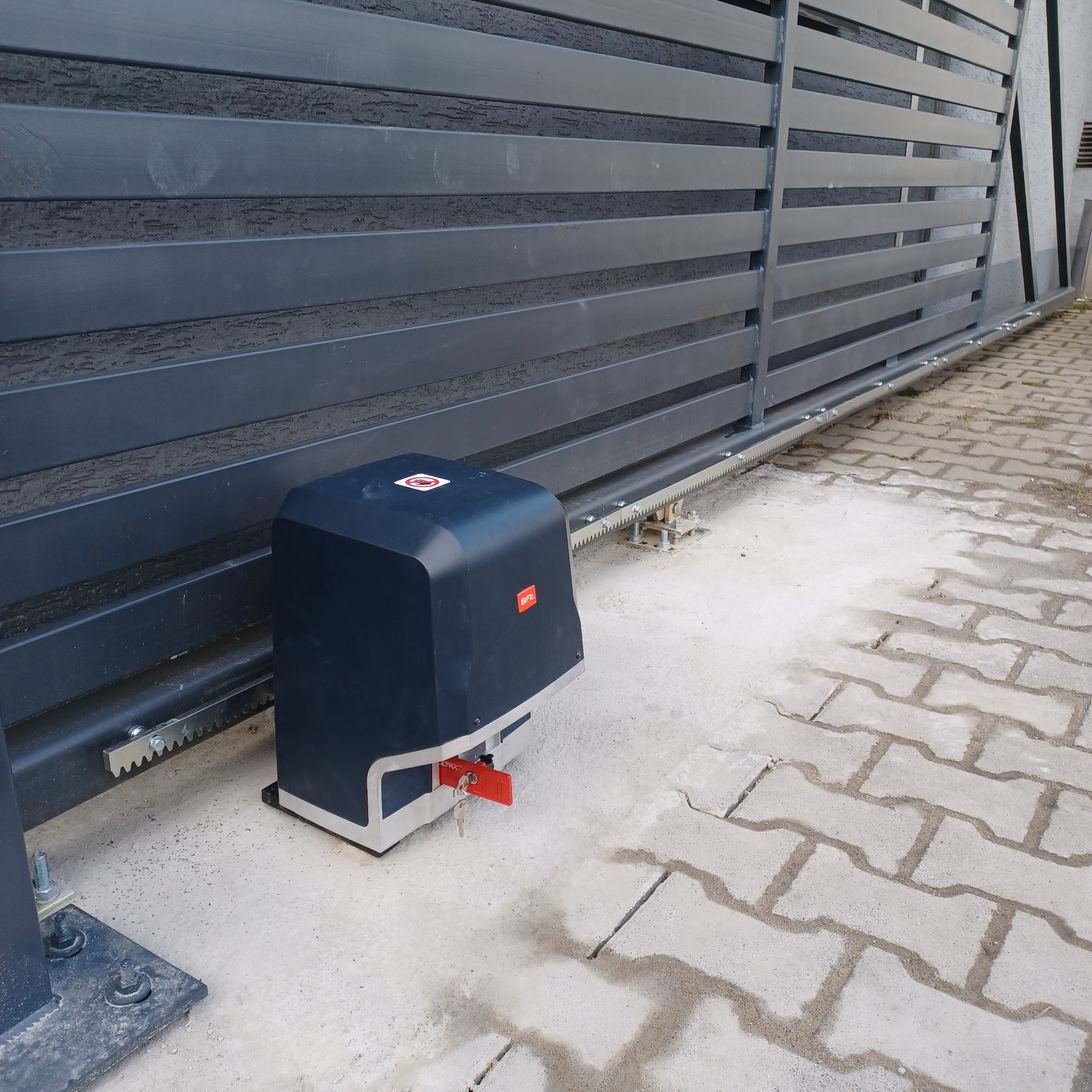 Zamontowany napęd do bramy przesuwnej z kluczykiem w Mosinie, widoczna listwa zębata i fragment bramy z poziomych profili.