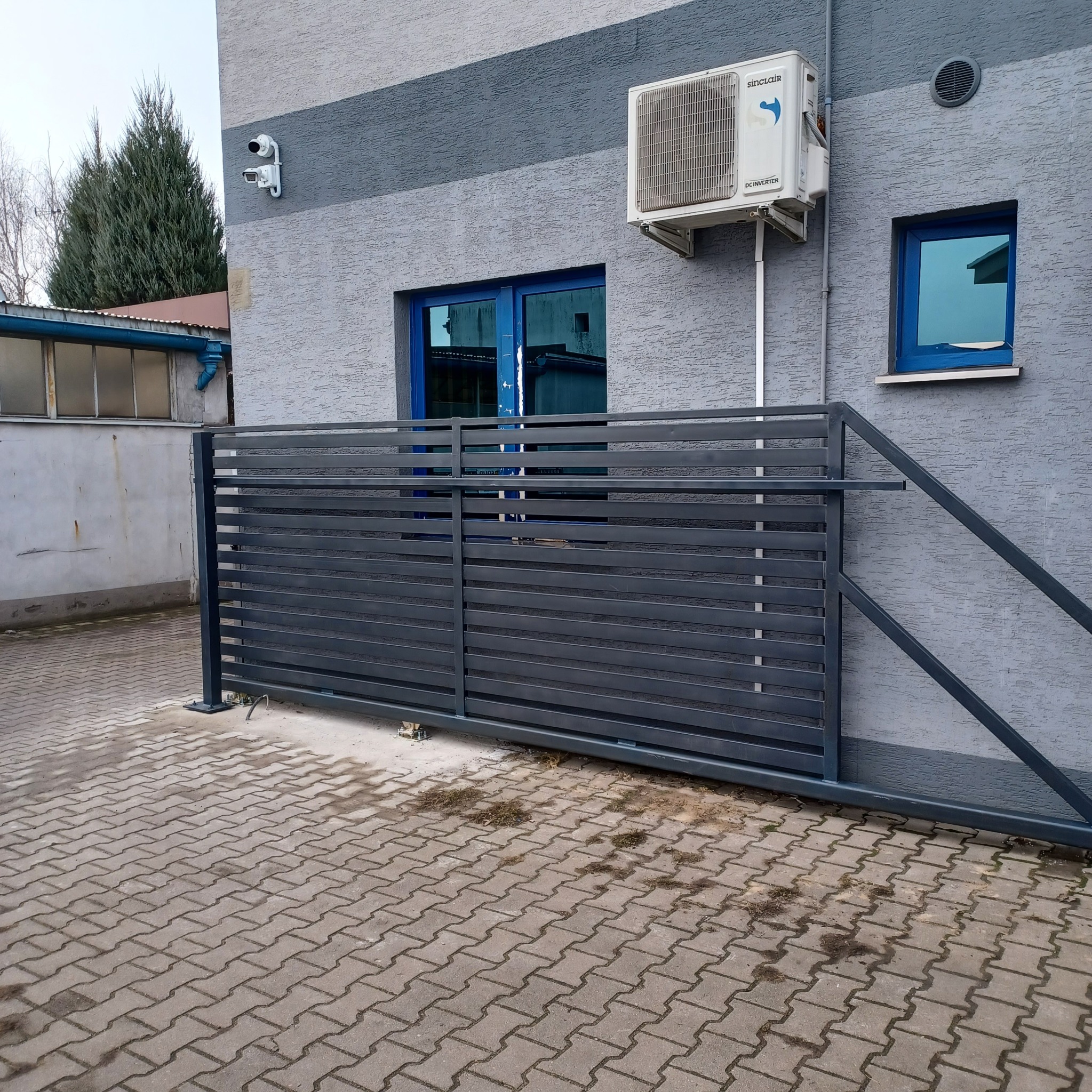 Nowoczesna brama przesuwna z poziomych profili stalowych w kolorze grafitowym, zamontowana przed budynkiem z szarą elewacją i niebieskimi oknami, widoczny fragment kostki brukowej.