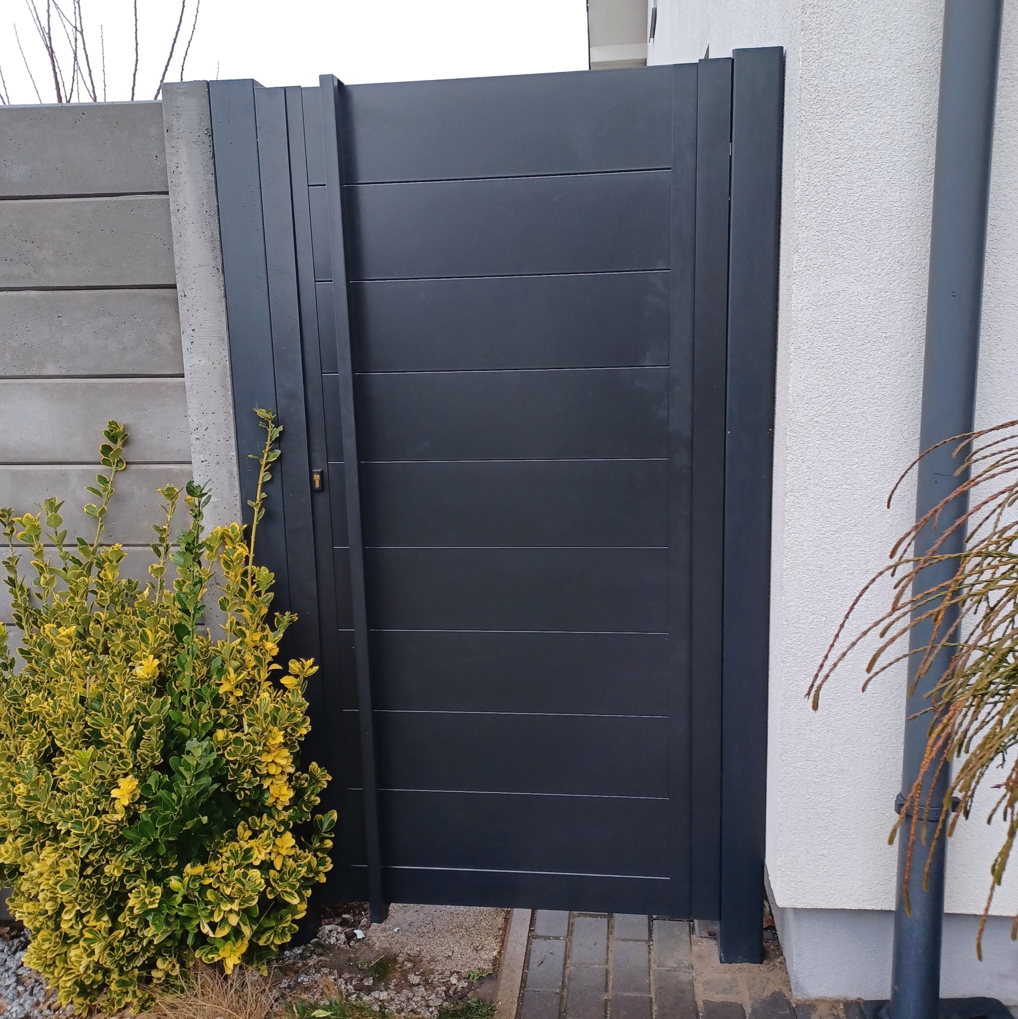 Nowoczesna, antracytowa furtka z poziomymi panelami, zamontowana przy betonowym ogrodzeniu i białej ścianie budynku, z widoczną roślinnością dekoracyjną.