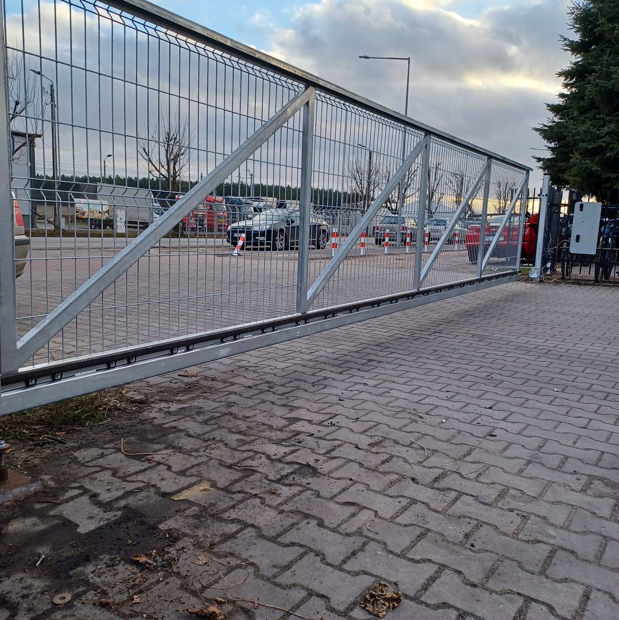 Przesuwna brama z metalowej siatki, widoczna od strony posesji, z samochodami i drzewami w tle, zamontowana na betonowej nawierzchni.