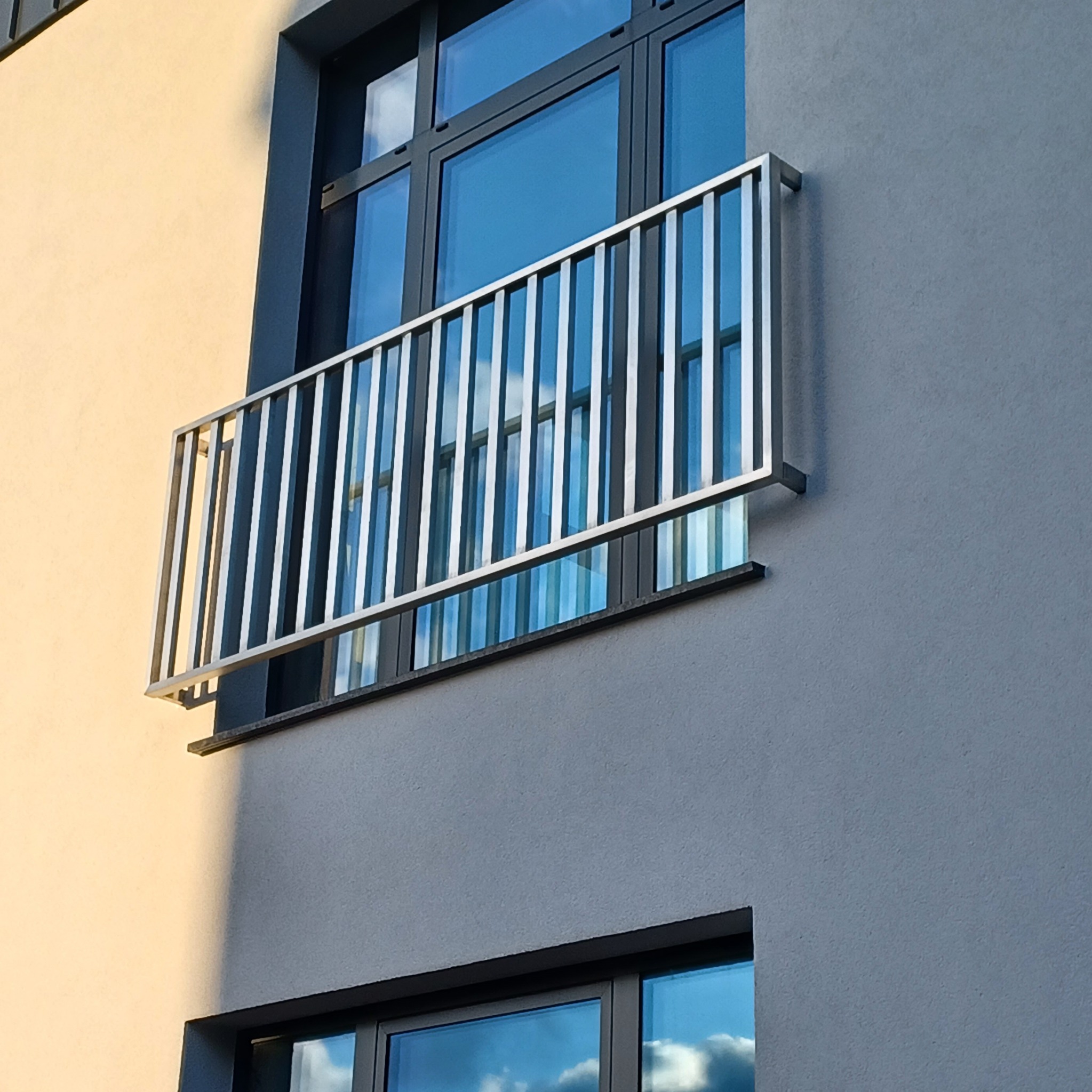 Nowoczesna balustrada balkonowa ze stali nierdzewnej, zamontowana na elewacji budynku z dużymi oknami, widok z dołu.